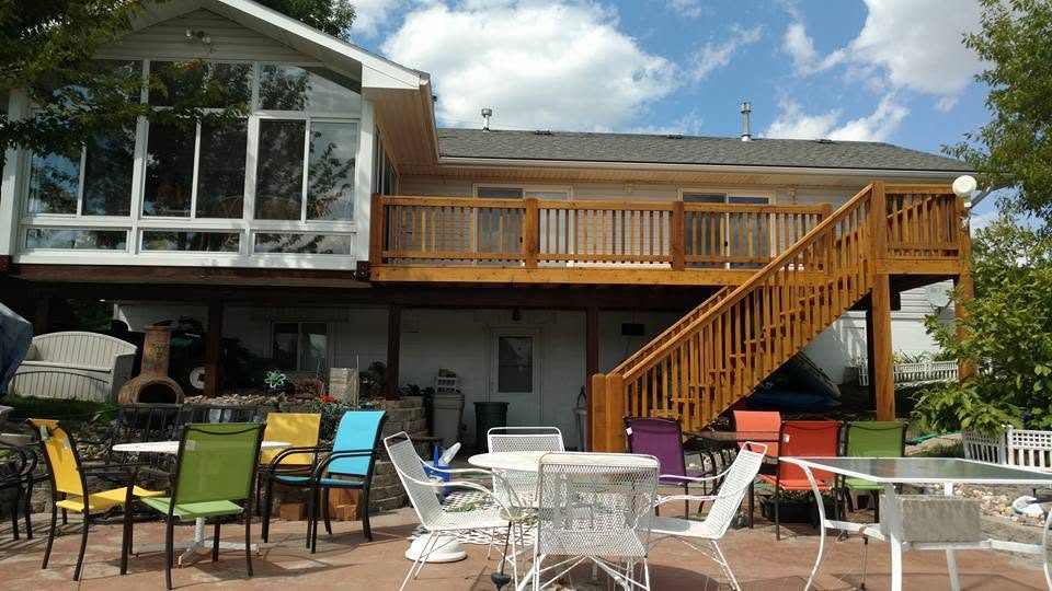 Residential deck with wooden railing, colorful patio chairs, and brick patio beneath two-story house