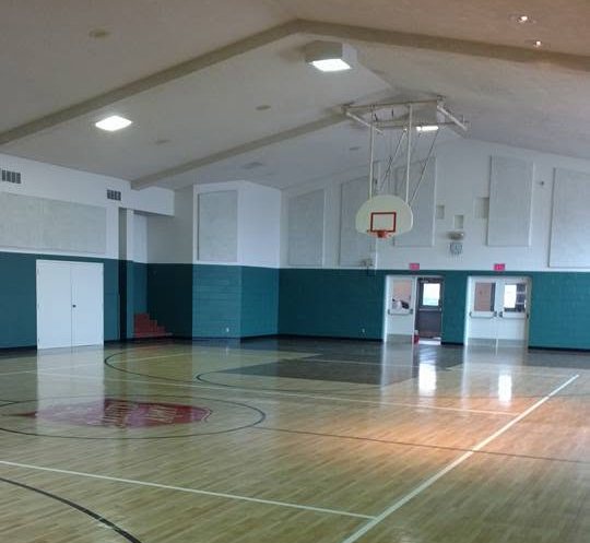 Interior of a school gymnasium with wooden flooring, basketball hoop, teal wall paneling, and fluorescent ceiling lights