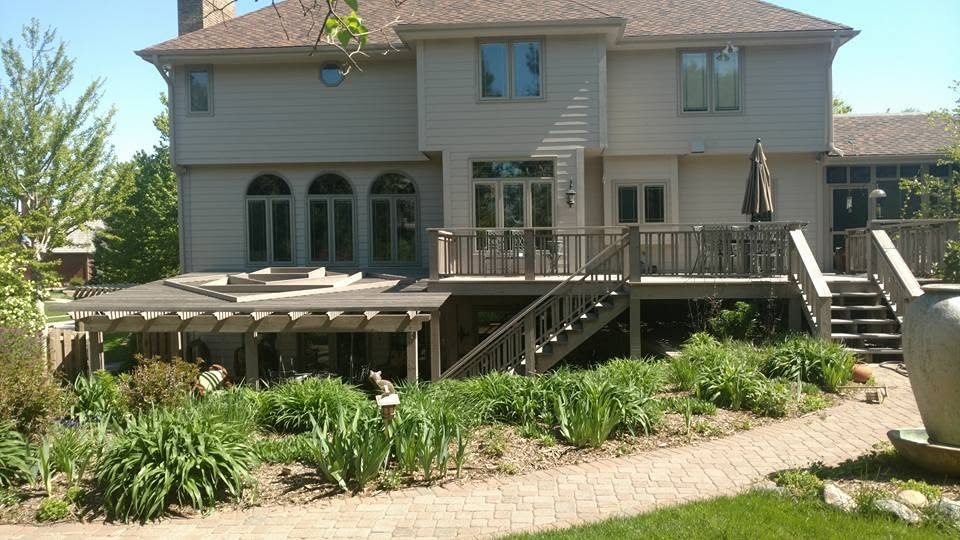 Two-story beige home with wooden deck, pergola, and brick patio in landscaped yard